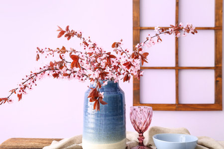 Vase with blooming tree branches, glass, bowl and napkin on wooden table near pink wall