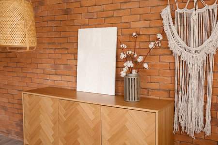 Stylish wooden cabinet with blooming cotton branches in vase near brown brick wall