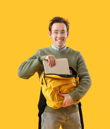 Male student putting laptop in backpack on yellow backgroundの写真素材