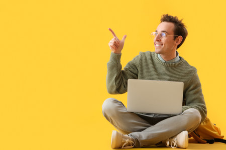 Male student with laptop pointing at something on yellow backgroundの写真素材