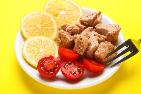 Plate with delicious canned tuna, fresh lemon, tomatoes and fork on yellow background, closeupの写真素材