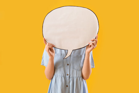 Young woman hiding face behind blank speech bubble on yellow backgroundの写真素材