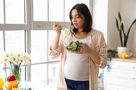 Young pregnant woman eating vegetable salad in the kitchenの写真素材