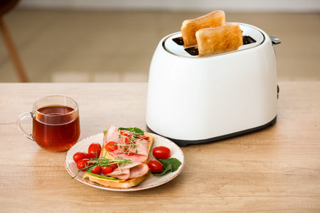 Modern toaster with crispy bread slices, tasty sandwiches and cup of tea on table in kitchen