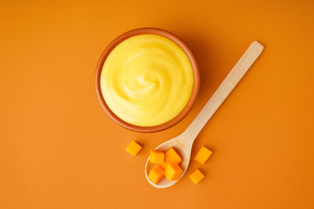 Bowl of tasty cheddar sauce and spoon with cheese cubes on orange backgroundの写真素材
