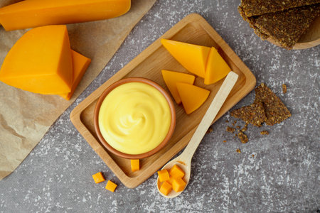 Board with bowl of tasty cheddar sauce and cheese chunks on black grunge backgroundの写真素材