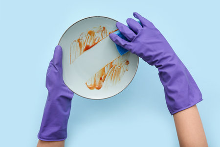 Female hands in rubber gloves washing dirty plate with sponge on blue backgroundの写真素材