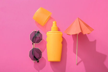Bottle of sunscreen cream with sunglasses and mini umbrella on pink background