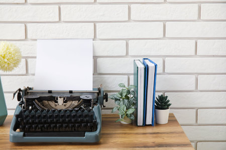 Vintage typewriter with vase of flowers, books and houseplants on brown wooden table near brick wallの写真素材