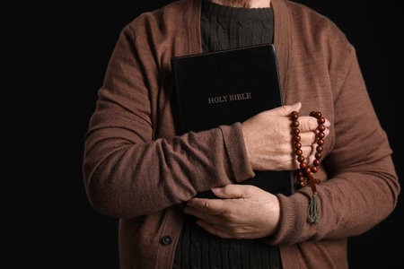 Senior woman with Holy Bible and prayer beads on black background, closeup