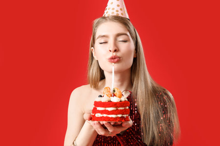 Young woman with birthday cake making wish on red background, closeup