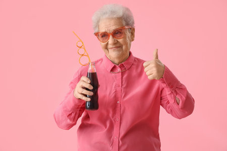 Senior woman with bottle of cola showing thumb-up on pink backgroundの写真素材