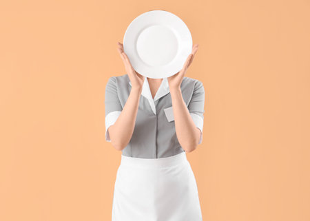 Young waitress with empty plate hiding face on beige backgroundの写真素材