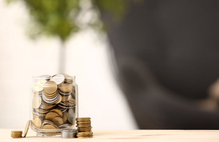 Jar with coins on wooden table against blurred background. Saving concept