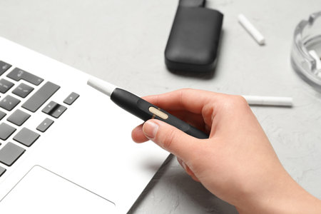 Woman holding modern electronic cigar near laptop on gray background