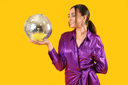 Smiling fashionable woman in elegant purple dress with disco ball posing on yellow backgroundの写真素材