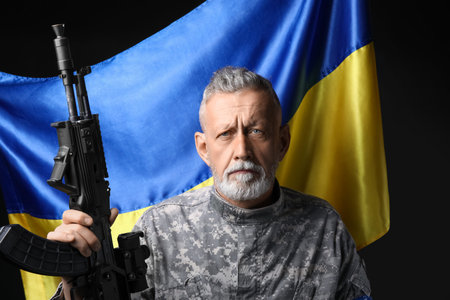 Mature soldier with assault rifle and flag of Ukraine on dark background