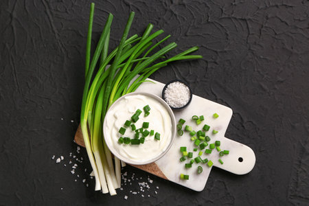 Board and bowl of tasty sour cream with sliced green onion on black backgroundの写真素材