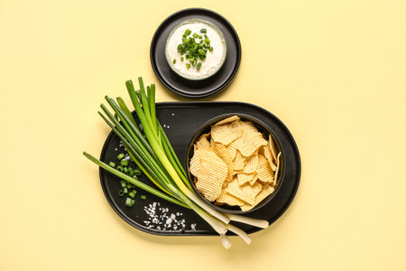 Bowl of tasty sour cream with sliced green onion and potato chips on yellow background