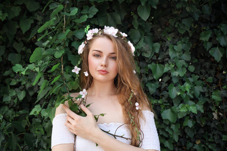 Beautiful young woman with flowers in her hair near green ivy outdoors. summer solsticeの写真素材