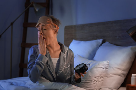 Sleepy young woman with alarm clock in bedroom at nightの写真素材