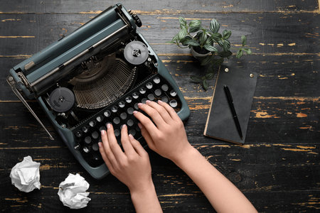 Woman typing on typewriter with notebook, pen and crumpled paper on dark wooden backgroundの写真素材