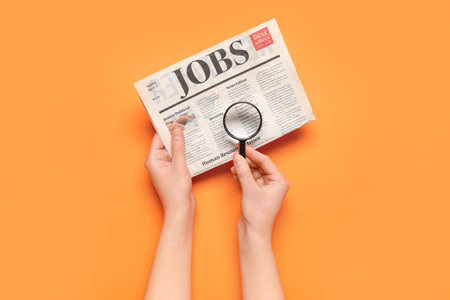 Female hands with mini magnifier and newspaper with headline JOBS on orange background. Search conceptの写真素材