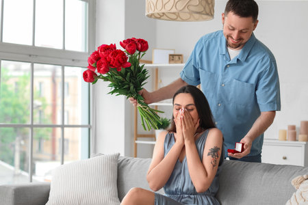 Young man with flowers proposing to his girlfriend at homeの写真素材