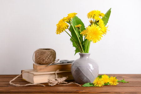 Vase with yellow dandelion flowers, rope, scissors and books on wooden table near light wallの写真素材