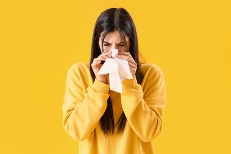 Allergic young woman with tissue sneezing on yellow backgroundの写真素材