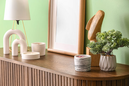 Holders with candles, blank frame, lamp and houseplant on chest of drawers near green wall in room