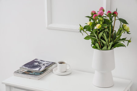 Vase with unopened peonies, cup of coffee and magazines on dresser near white wallの写真素材