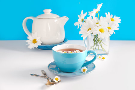 Teapot with cup of natural chamomile tea and flowers on white table near blue wallの写真素材