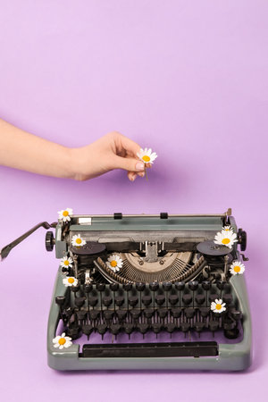 Woman putting daisy on vintage typewriter on lilac backgroundの写真素材