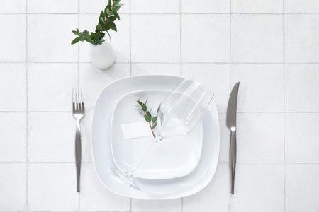 Stylish table setting with glass and blank card on white tile background