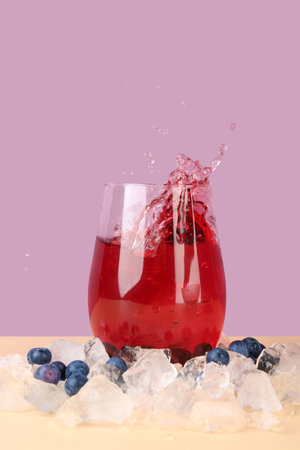 Glass of tasty blueberry cider with splashes, ice cubes and berries on beige tableの写真素材