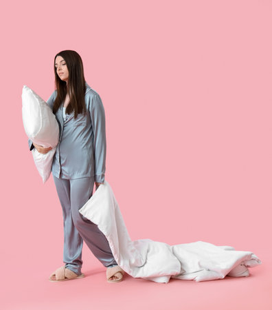 Young woman in pajamas with blanket and pillow on pink background