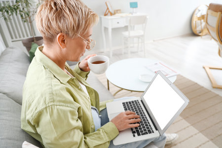 Mature female programmer working with laptop and cup of tea on sofa at home, closeupの写真素材
