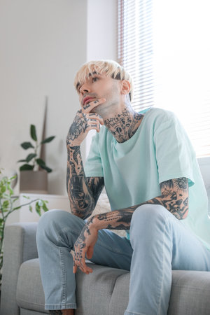 Thoughtful young tattooed man sitting on sofa at homeの写真素材