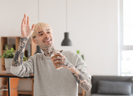 Young tattooed man with cup of coffee waving hand at homeの写真素材
