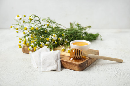 Wooden board with bowl of honey, tea bags and fresh chamomile flowers on light backgroundの写真素材
