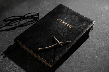 Holy Bible with wooden cross and eyeglasses on black background