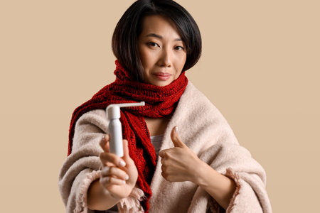 Ill Asian woman with spray for sore throat showing thumb-up on beige backgroundの写真素材