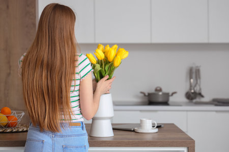 Beautiful young woman putting bouquet of yellow tulip flowers into vase in modern kitchenの写真素材
