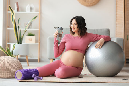 Sporty pregnant woman with bottle of water and fitball at homeの写真素材
