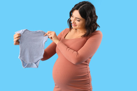 Young pregnant woman with baby bodysuit on blue backgroundの写真素材