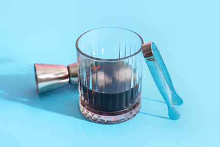 Glass of rum with ice cubes, tongs and measure cup on blue background, closeupの写真素材