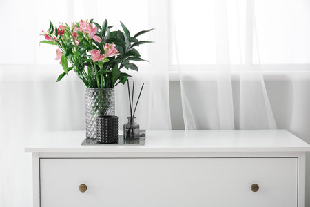 Reed diffuser and vase with beautiful flowers on chest of drawers near windowの写真素材