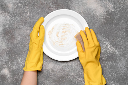 Female hands in rubber gloves washing dirty plate on gray grunge backgroundの写真素材