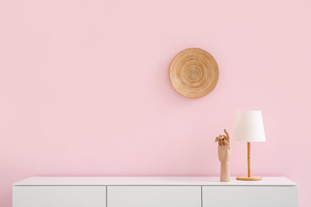 Modern chest of drawers with lamp, wooden hand and plate hanging on pink wall in roomの写真素材